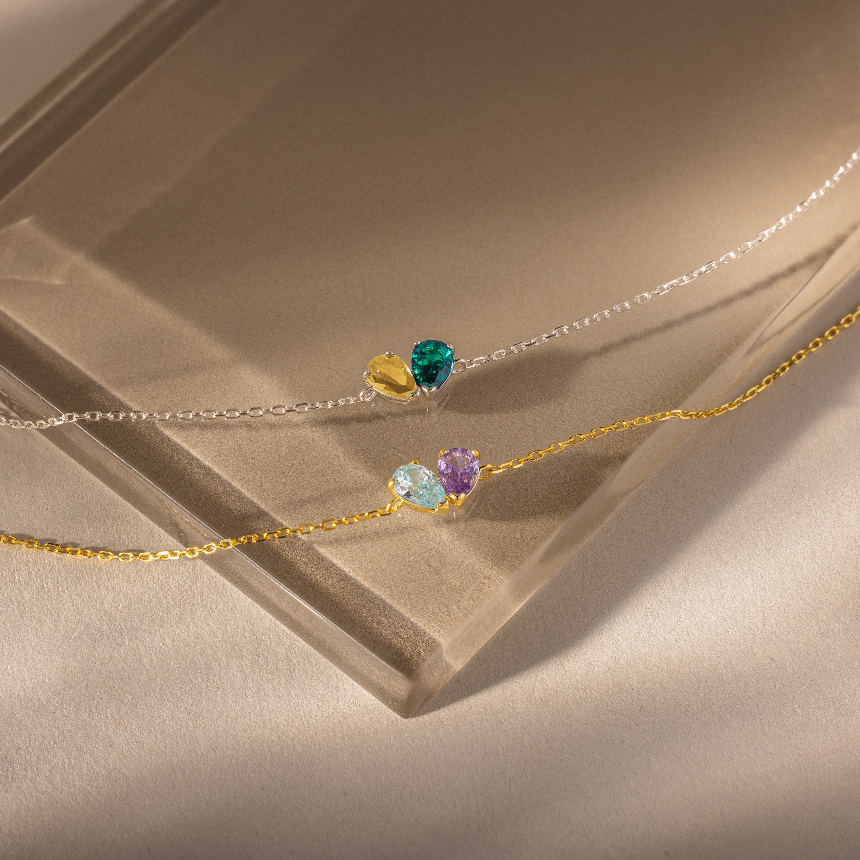 The Toi et Moi Teardrop Heart Bracelet in Sterling Silver, featuring colorful gemstone heart charms, rests alongside another delicate chain bracelet on a sunlit, neutral surface.