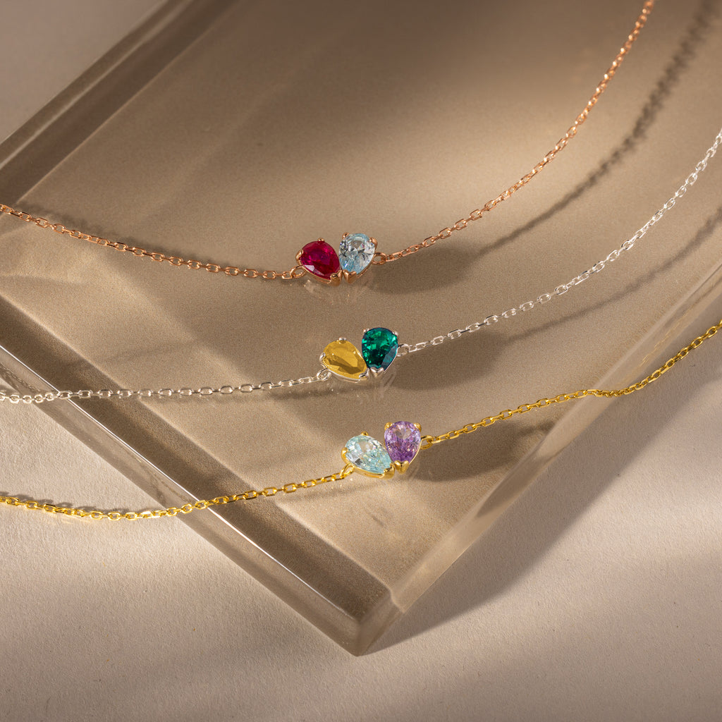 The Toi et Moi Teardrop Heart Bracelet features delicate chains and colorful teardrop heart pendants, elegantly displayed on a beige glass surface.