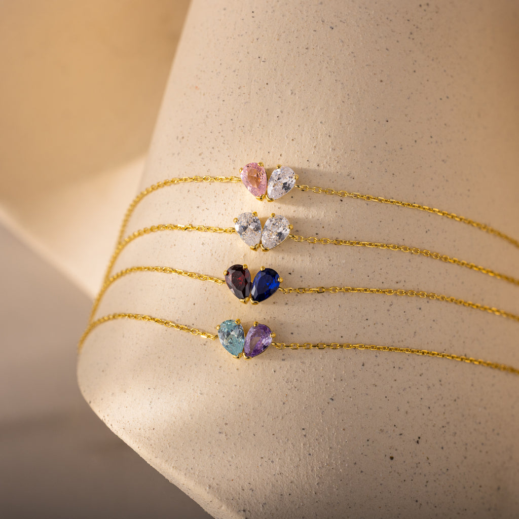 Four Toi et Moi Teardrop Heart Bracelets in 18K Gold, featuring colorful pear-shaped and dual gemstones, displayed on a beige textured stand.