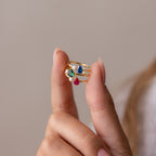 A hand displays three gold rings, including a Teardrop Pink Tourmaline & Pearl Ring with colorful gemstones, against a neutral background for an elegant jewelry showcase.