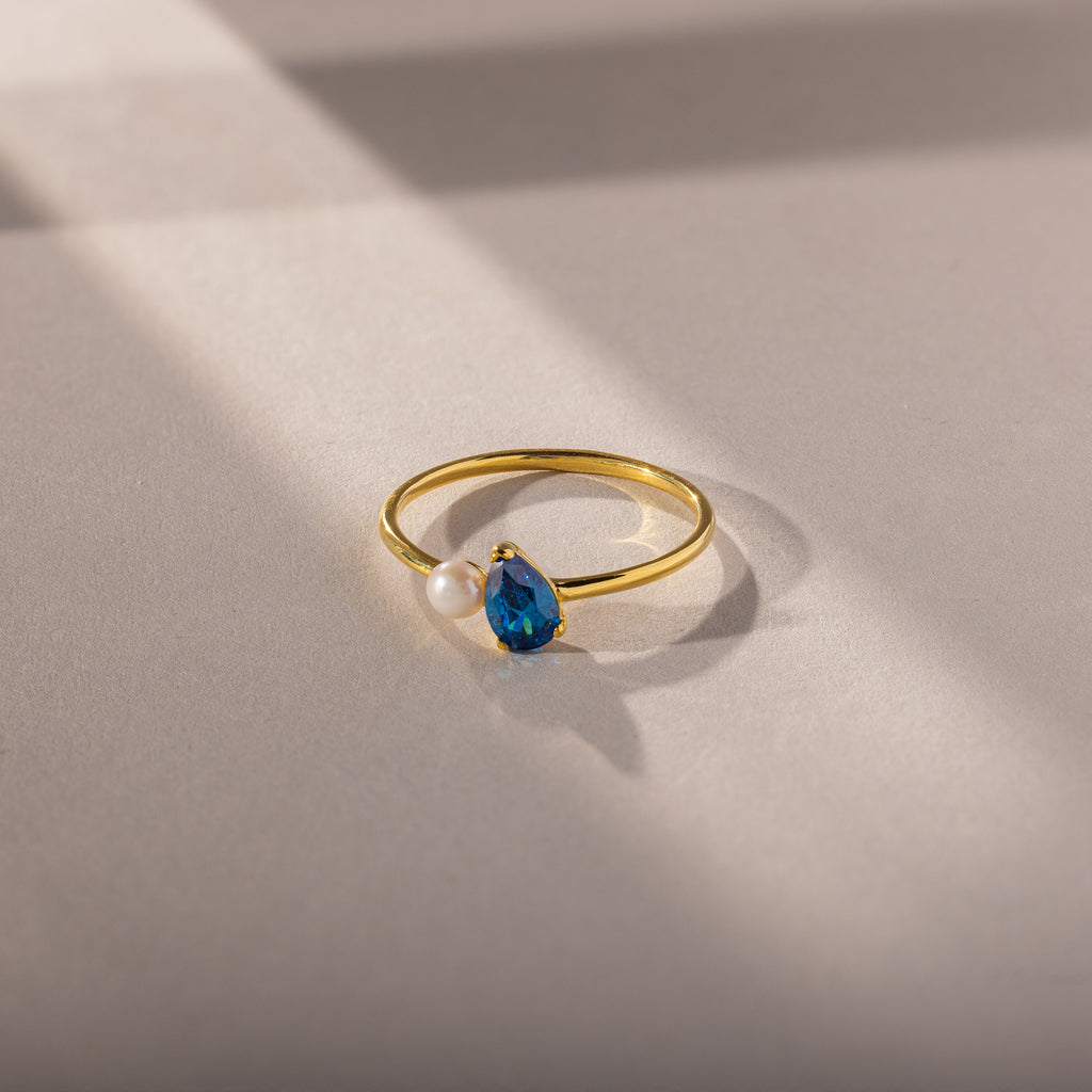 The Teardrop Blue Zircon & Pearl Ring features a blue zircon and pearl, elegantly displayed on a softly lit beige surface with delicate shadow lines—an exquisite piece of jewelry.