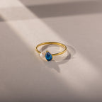 The Teardrop Blue Zircon & Pearl Ring features a blue zircon and pearl, elegantly displayed on a softly lit beige surface with delicate shadow lines—an exquisite piece of jewelry.