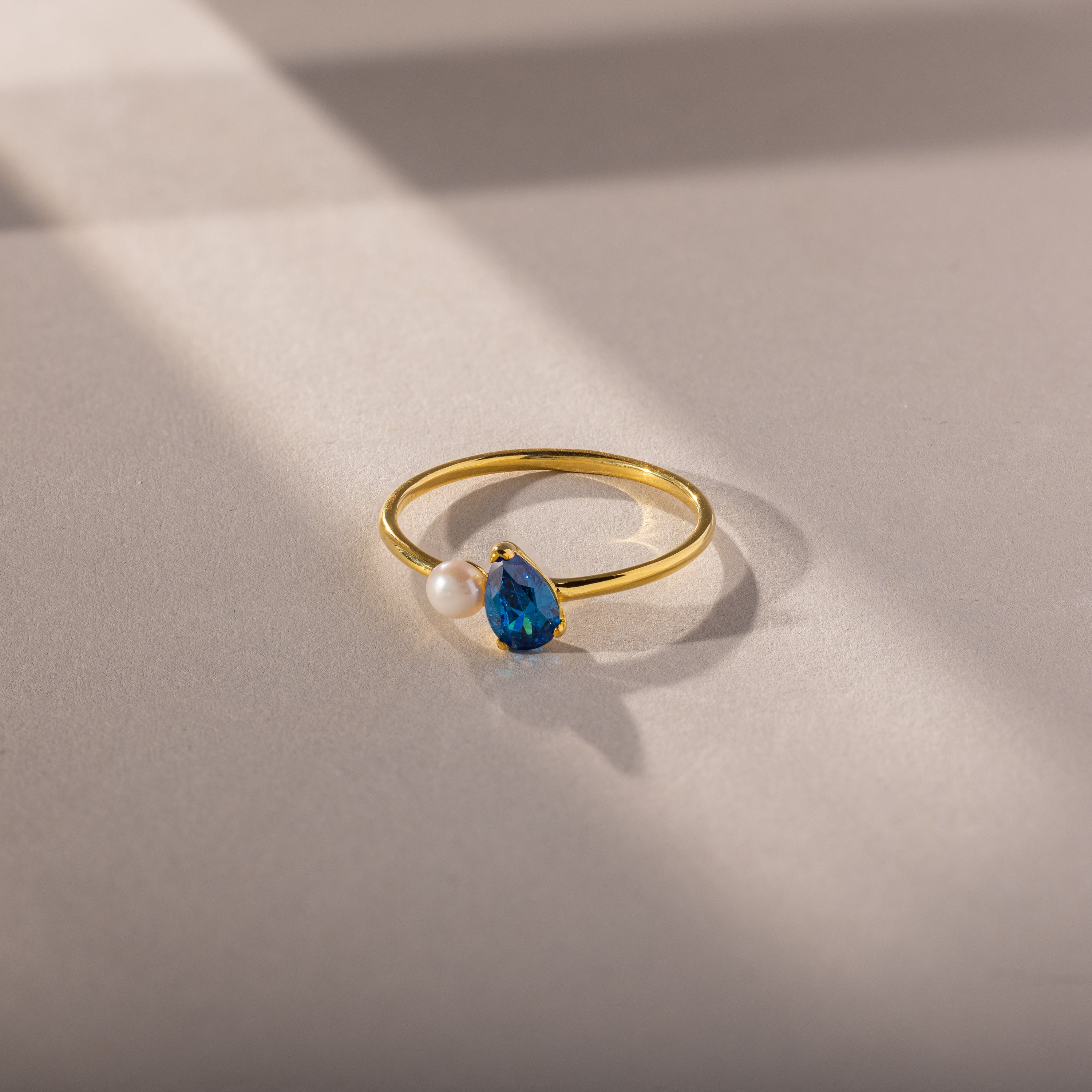 The Teardrop Blue Zircon & Pearl Ring features a blue zircon and pearl, elegantly displayed on a softly lit beige surface with delicate shadow lines—an exquisite piece of jewelry.