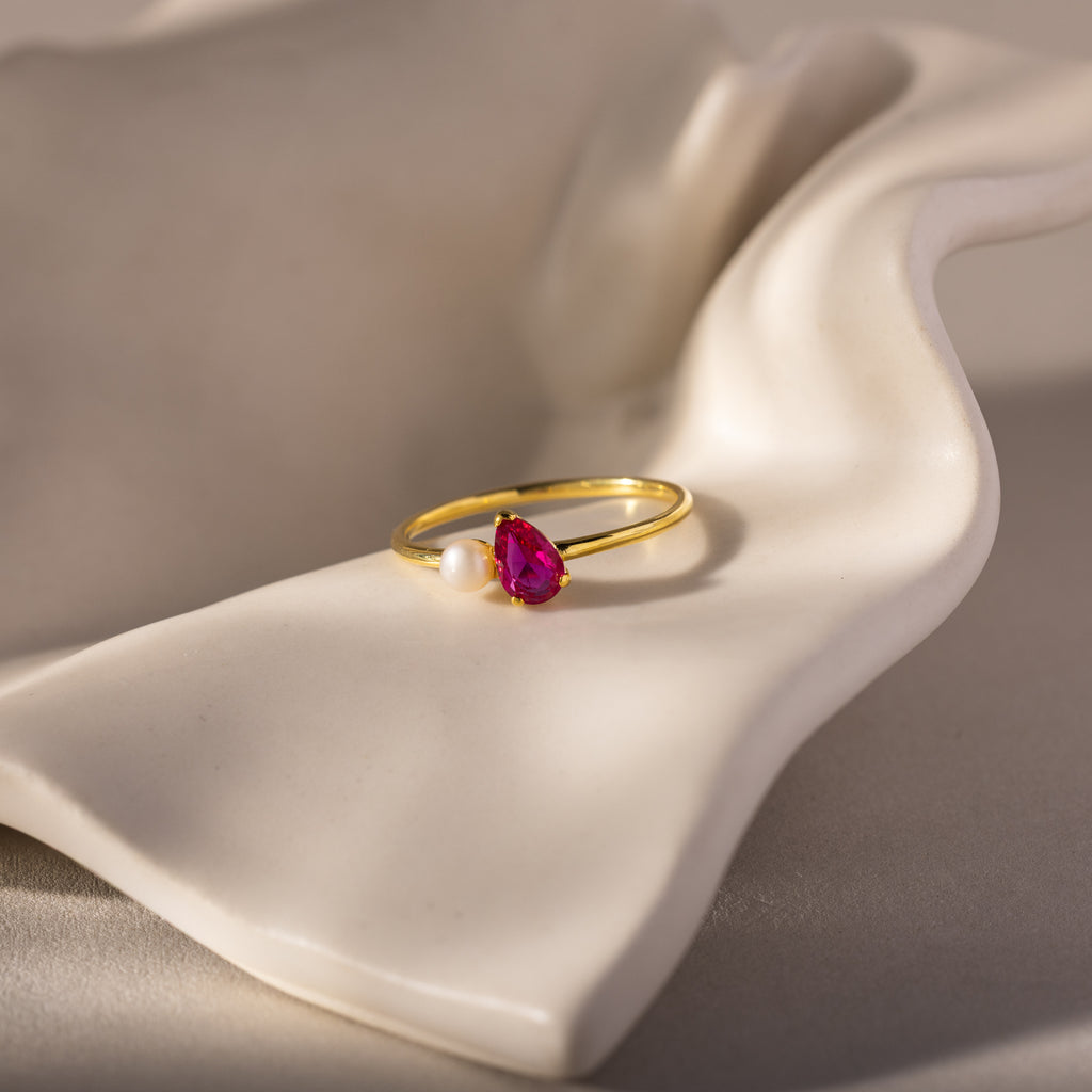 The Teardrop Ruby & Pearl Ring, featuring a radiant red gemstone and a lustrous pearl, rests on a white, wavy ceramic surface—a true symbol of elegance.