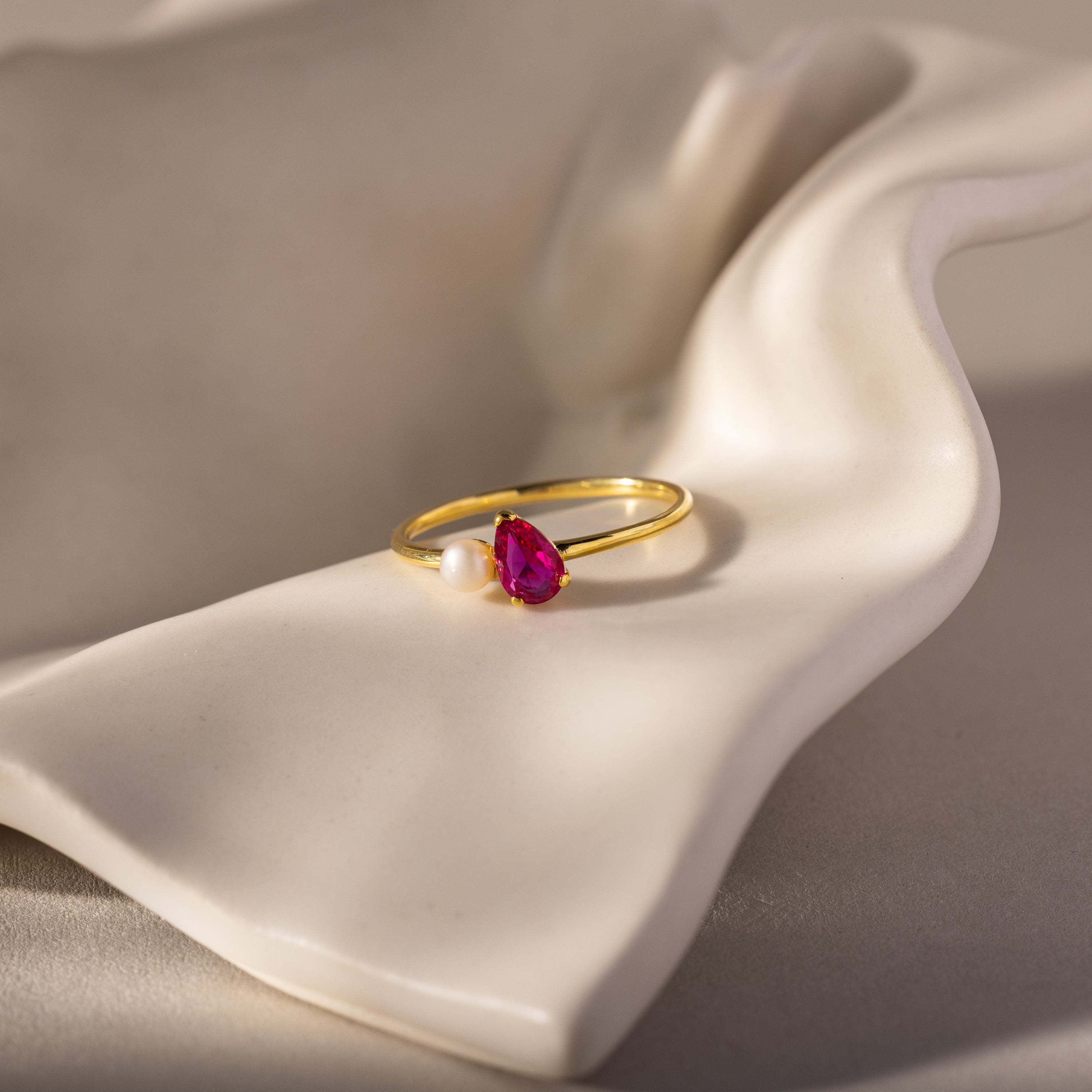 The Teardrop Ruby & Pearl Ring, featuring a radiant red gemstone and a lustrous pearl, rests on a white, wavy ceramic surface—a true symbol of elegance.