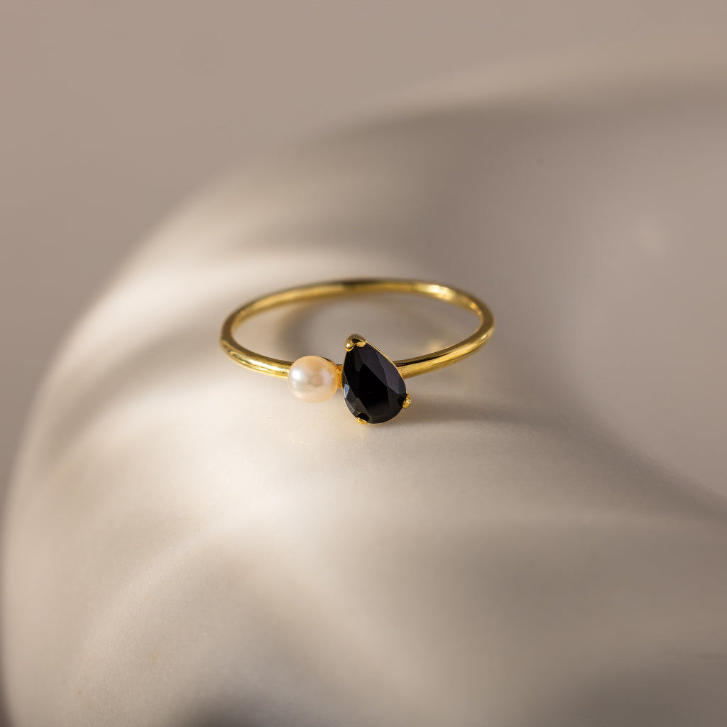 The Teardrop Birthstone & Pearl Ring features a black teardrop birthstone paired with a petite white pearl, elegantly displayed on a smooth, light surface.