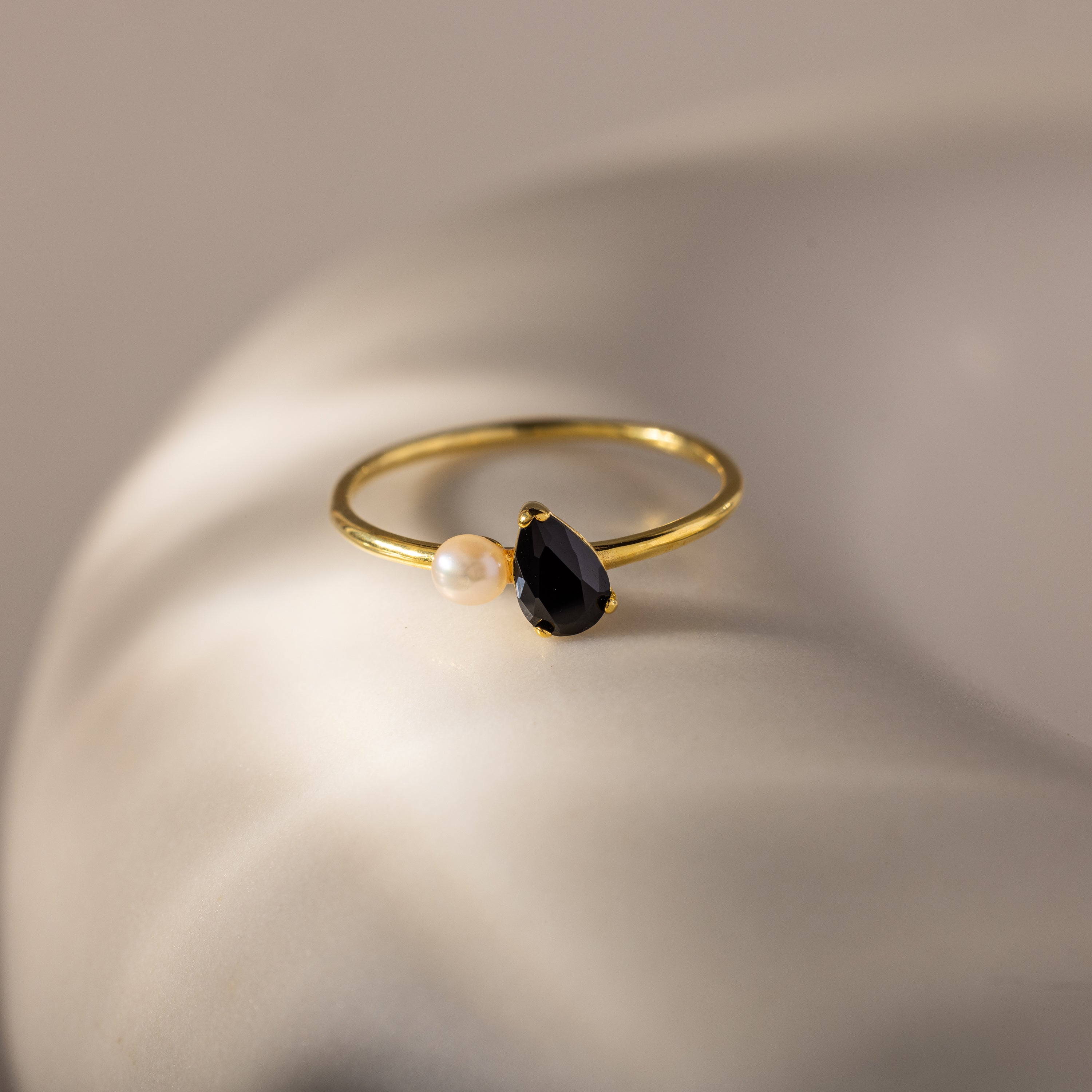 The Teardrop Birthstone & Pearl Ring features a black teardrop birthstone paired with a petite white pearl, elegantly displayed on a smooth, light surface.