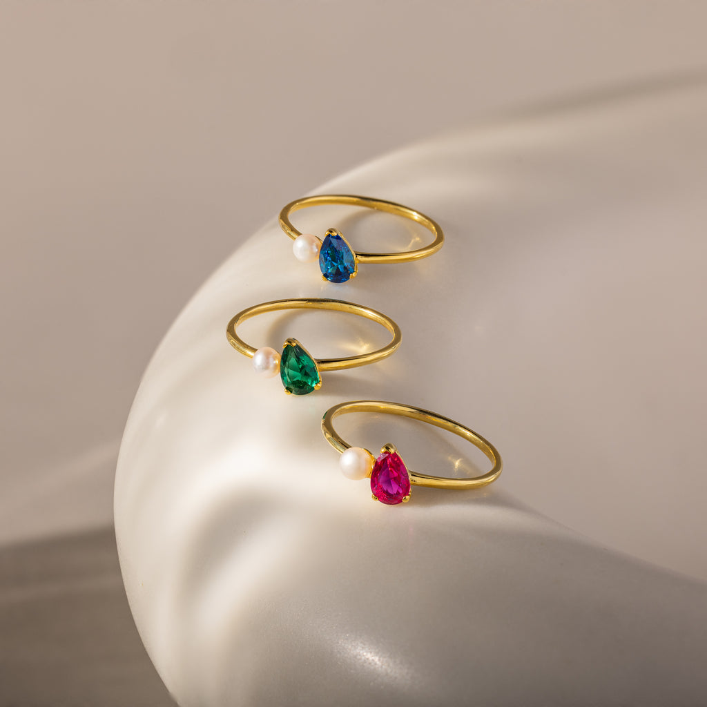 The Teardrop Birthstone & Pearl Ring features colored gems and a classic pearl, beautifully displayed on a smooth white surface—ideal for elegant jewelry lovers.