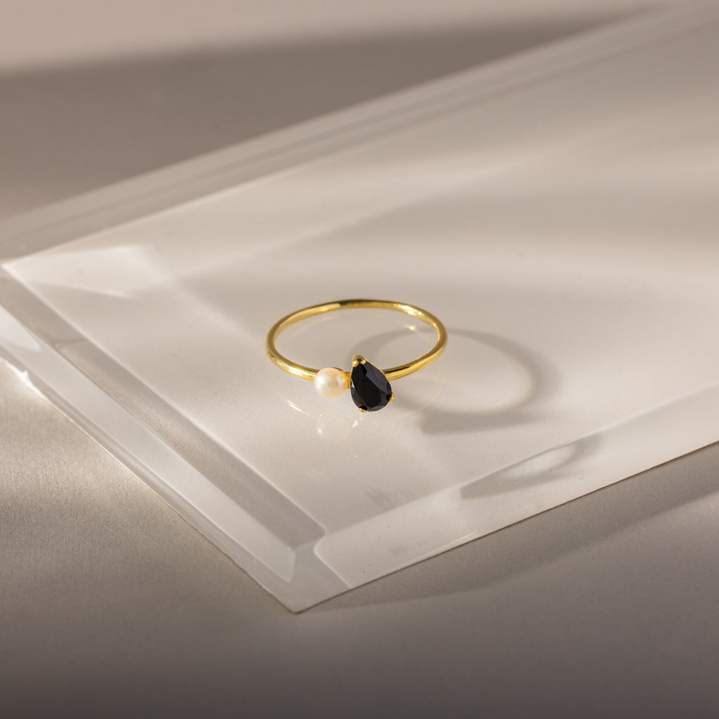 The Teardrop Onyx & Pearl Ring is an elegant gold piece featuring a black onyx teardrop birthstone and a white pearl set on a clear, angled surface.