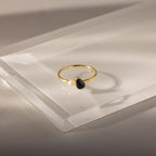 The Teardrop Onyx & Pearl Ring is an elegant gold piece featuring a black onyx teardrop birthstone and a white pearl set on a clear, angled surface.