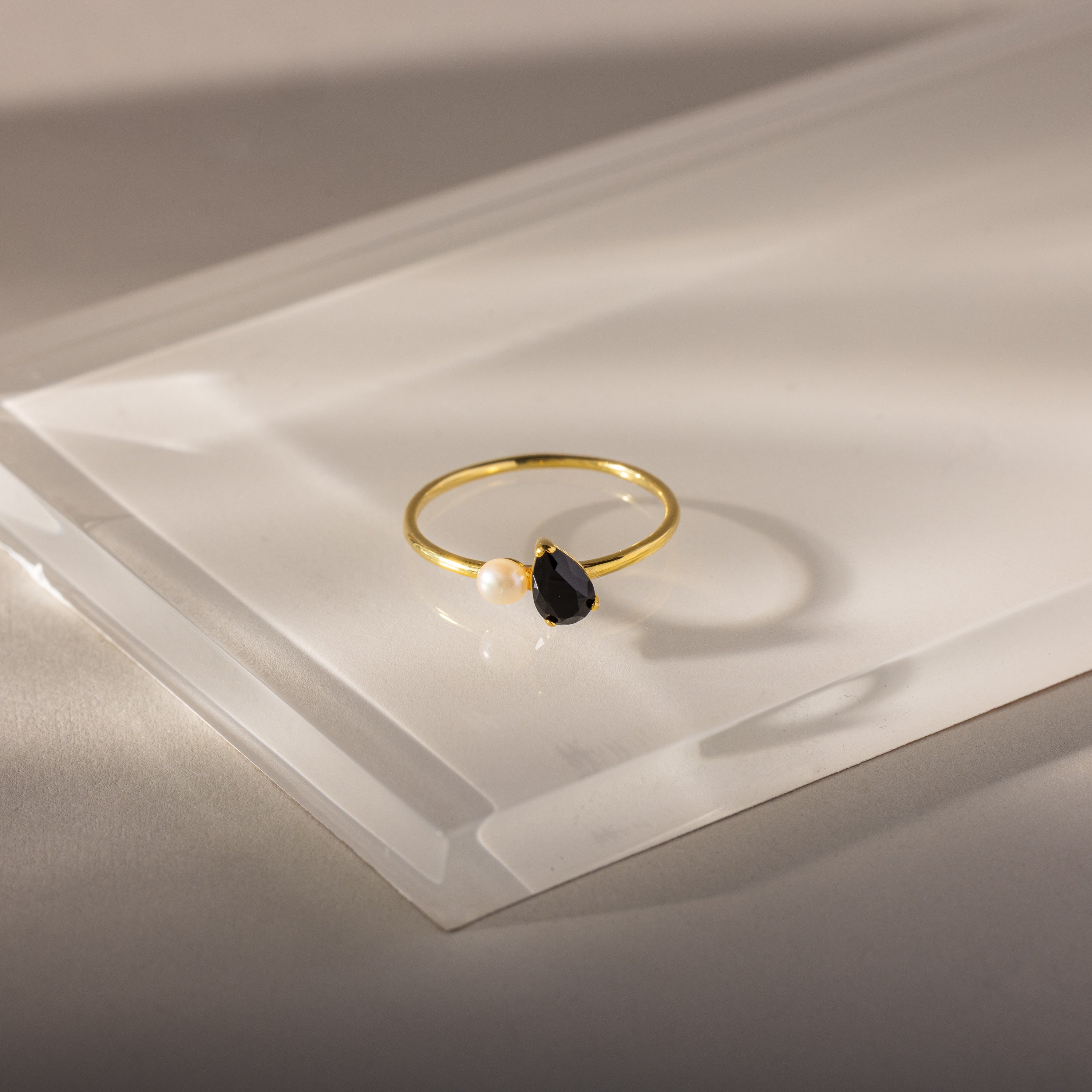 The Teardrop Onyx & Pearl Ring is an elegant gold piece featuring a black onyx teardrop birthstone and a white pearl set on a clear, angled surface.