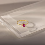 The Teardrop Ruby & Pearl Ring showcases a vivid red teardrop stone and gold pearls, elegantly displayed on a clear acrylic surface.