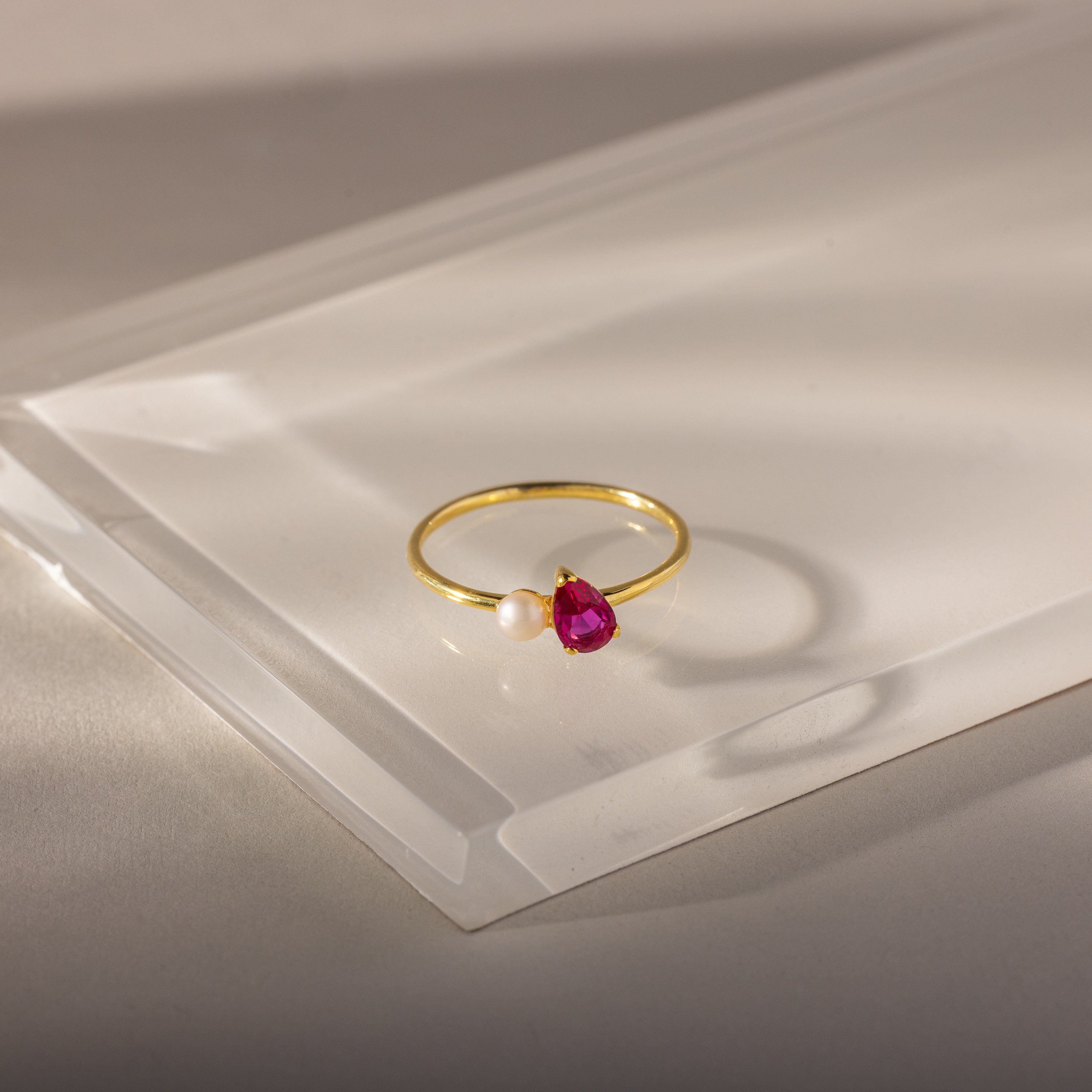 The Teardrop Ruby & Pearl Ring showcases a vivid red teardrop stone and gold pearls, elegantly displayed on a clear acrylic surface.