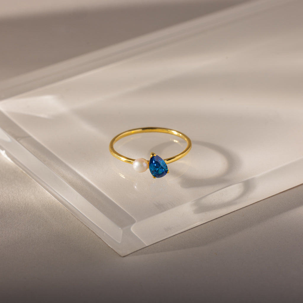 Teardrop Blue Zircon & Pearl Ring, featuring a blue zircon gem and a white pearl in an elegant design, displayed on clear acrylic.