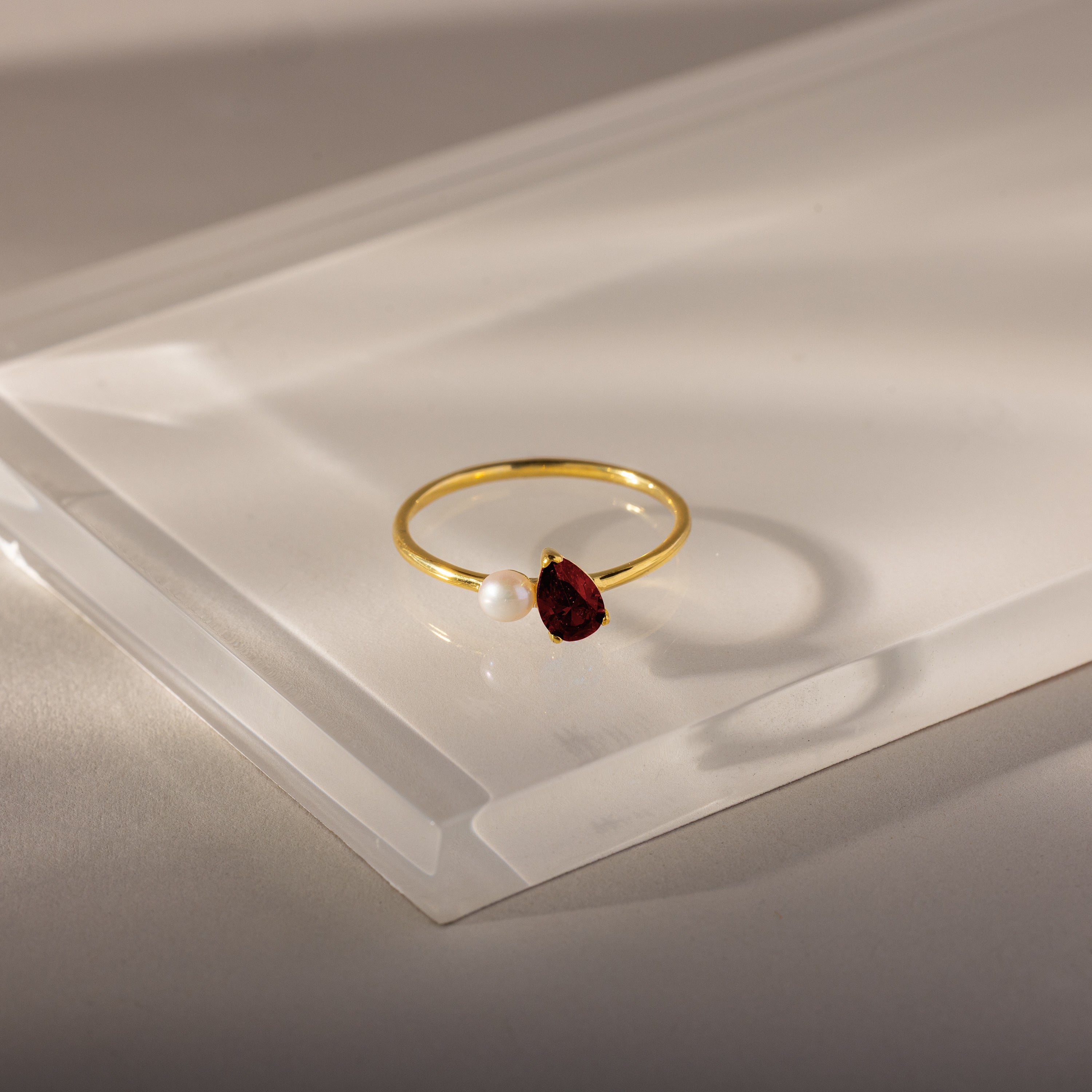 The Teardrop Garnet & Pearl Ring, featuring a red garnet and a small white pearl in a gold setting, rests on a clear rectangular surface, highlighting its elegant jewelry design.