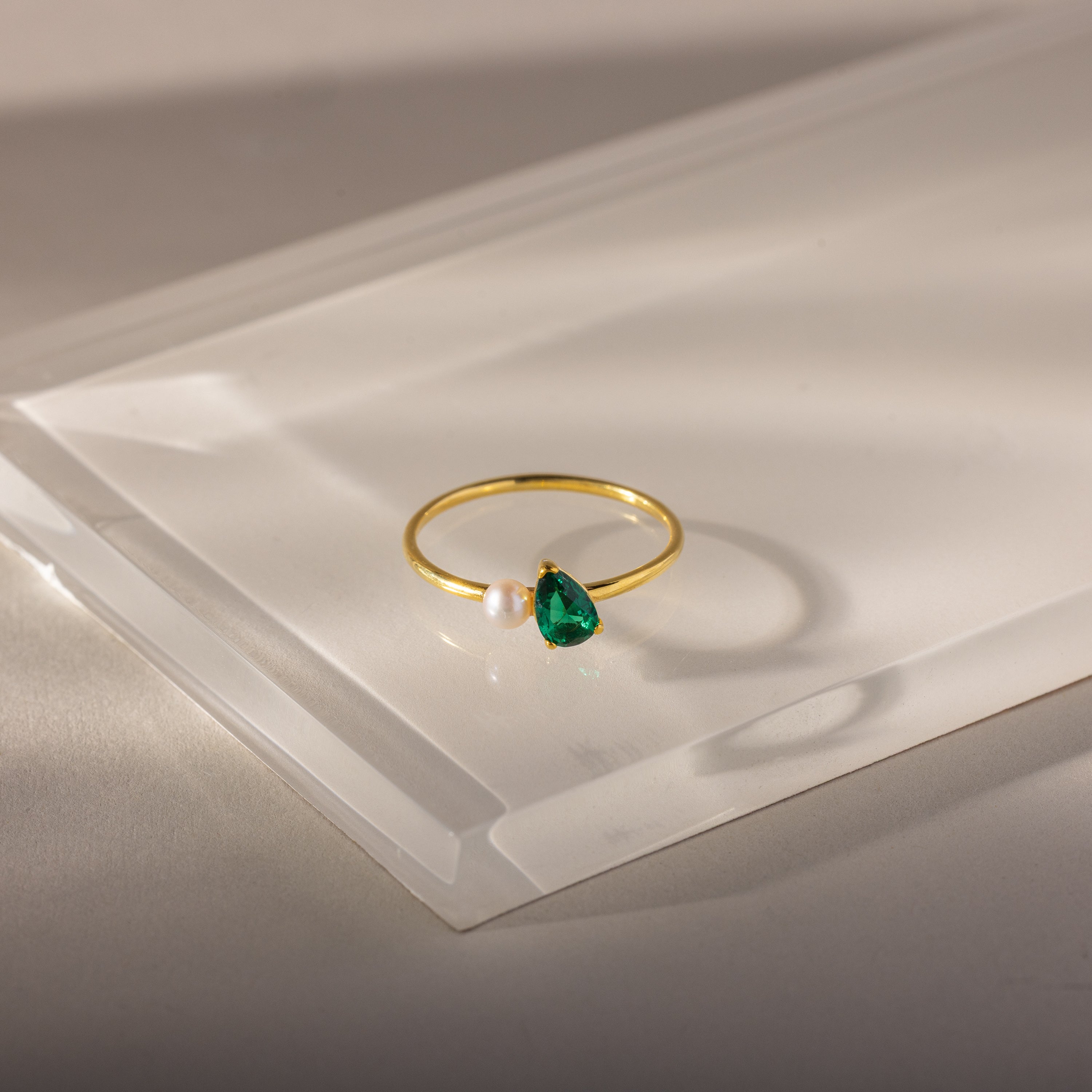 The Teardrop Emerald & Pearl Ring is an elegant gold jewelry piece showcasing a green gem and a small white pearl, beautifully displayed on a clear acrylic surface.