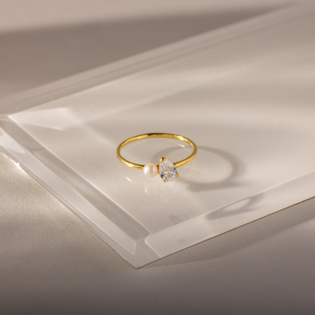 The Teardrop Opal & Pearl Ring, featuring a radiant pearl and opal gemstone, rests on a clear angled acrylic surface—a stunning piece of elegant jewelry.