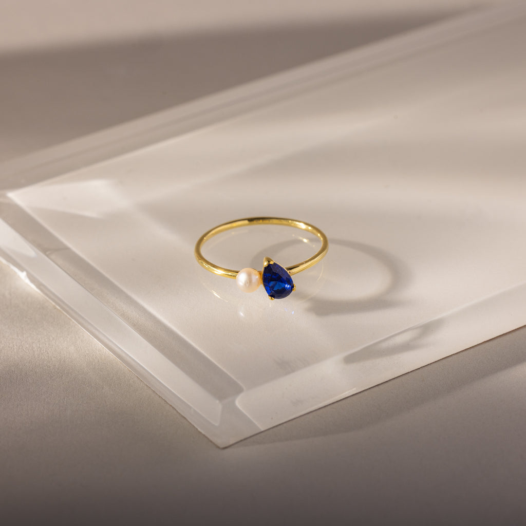 The Teardrop Sapphire & Pearl Ring features a gold band with a teardrop-shaped blue sapphire and a delicate pearl, displayed on a clear, angled surface—an elegant and exquisite jewelry piece.