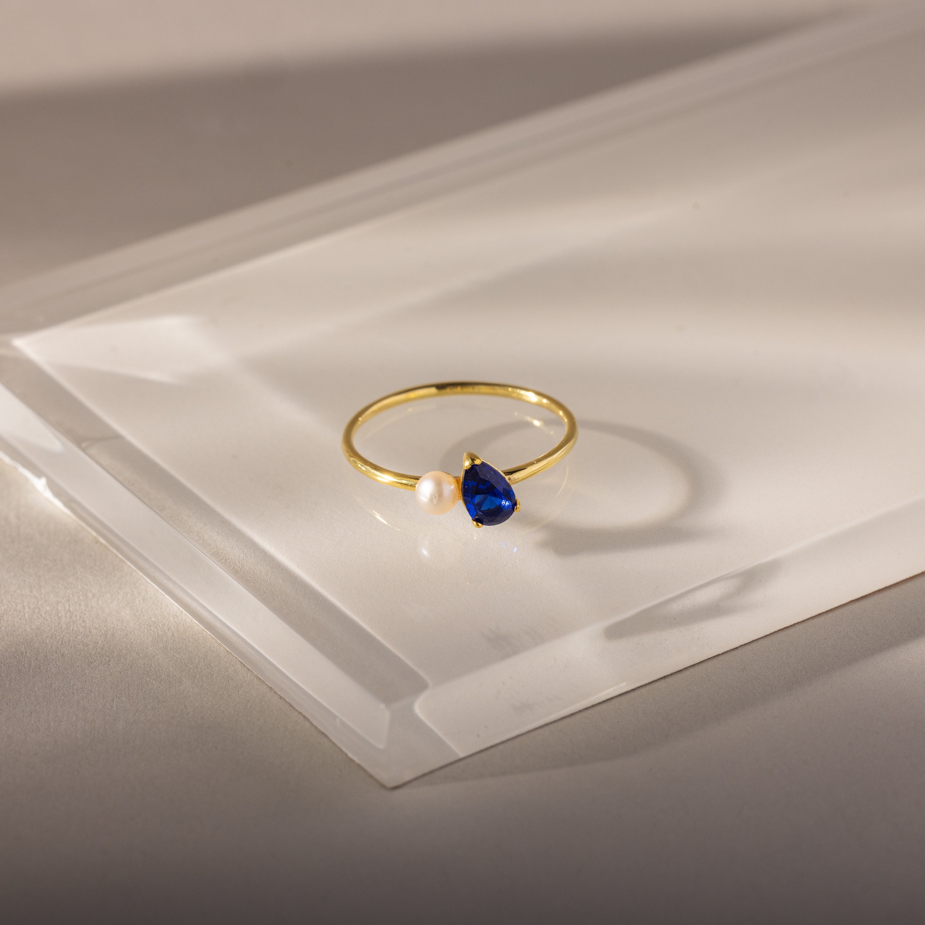 The Teardrop Sapphire & Pearl Ring features a gold band with a teardrop-shaped blue sapphire and a delicate pearl, displayed on a clear, angled surface—an elegant and exquisite jewelry piece.