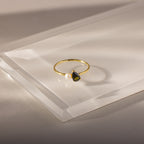 The Teardrop Peridot & Pearl Ring, featuring a teardrop peridot birthstone and a delicate white pearl, is displayed on clear acrylic—an elegant and exquisite jewelry piece.