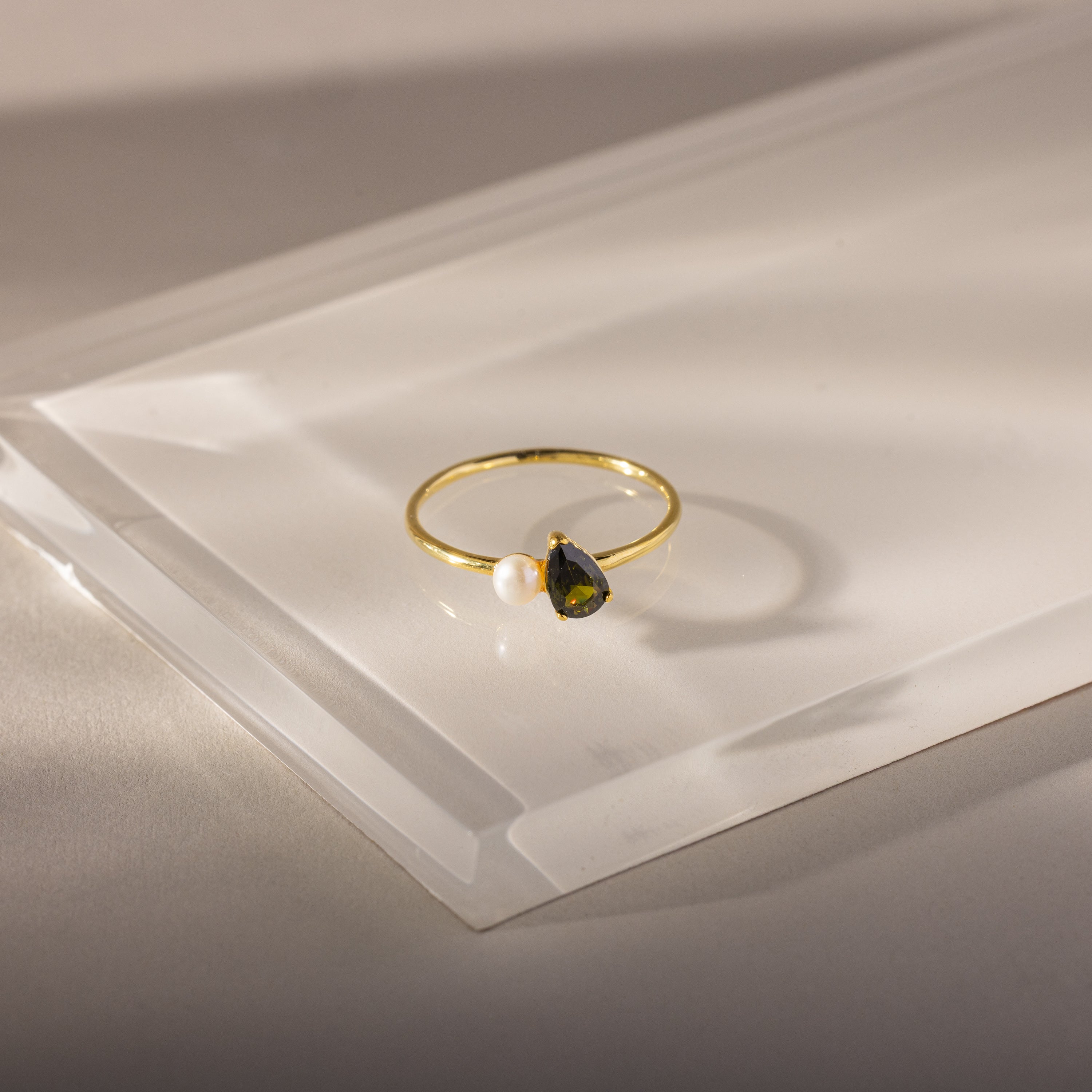 The Teardrop Peridot & Pearl Ring, featuring a teardrop peridot birthstone and a delicate white pearl, is displayed on clear acrylic—an elegant and exquisite jewelry piece.