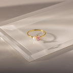 The Teardrop Pink Tourmaline & Pearl Ring, featuring a pink tourmaline stone and a delicate white pearl, rests atop a clear rectangular surface—an elegant jewelry piece with lasting charm.