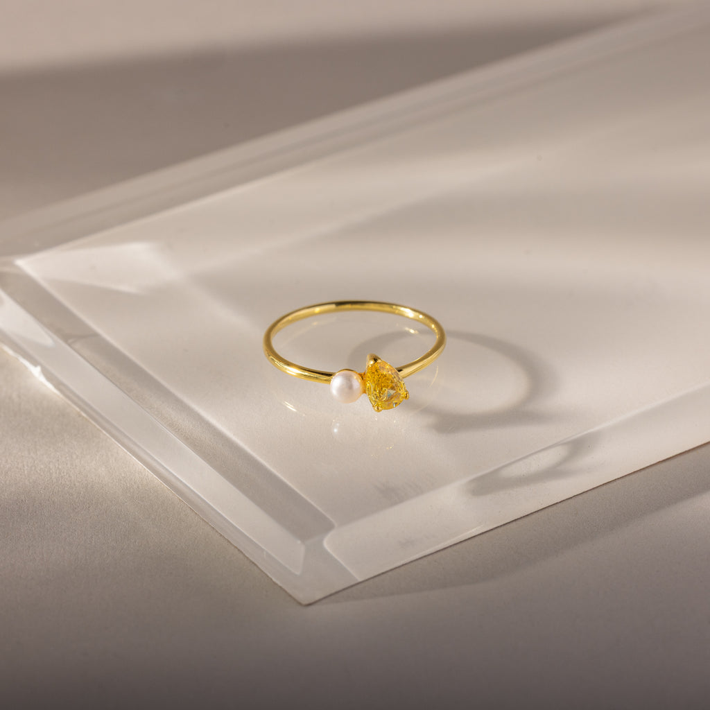 The Teardrop Topaz & Pearl Ring, featuring a yellow topaz and a white pearl, sits on a translucent surface, highlighting elegant jewelry perfect for any occasion.