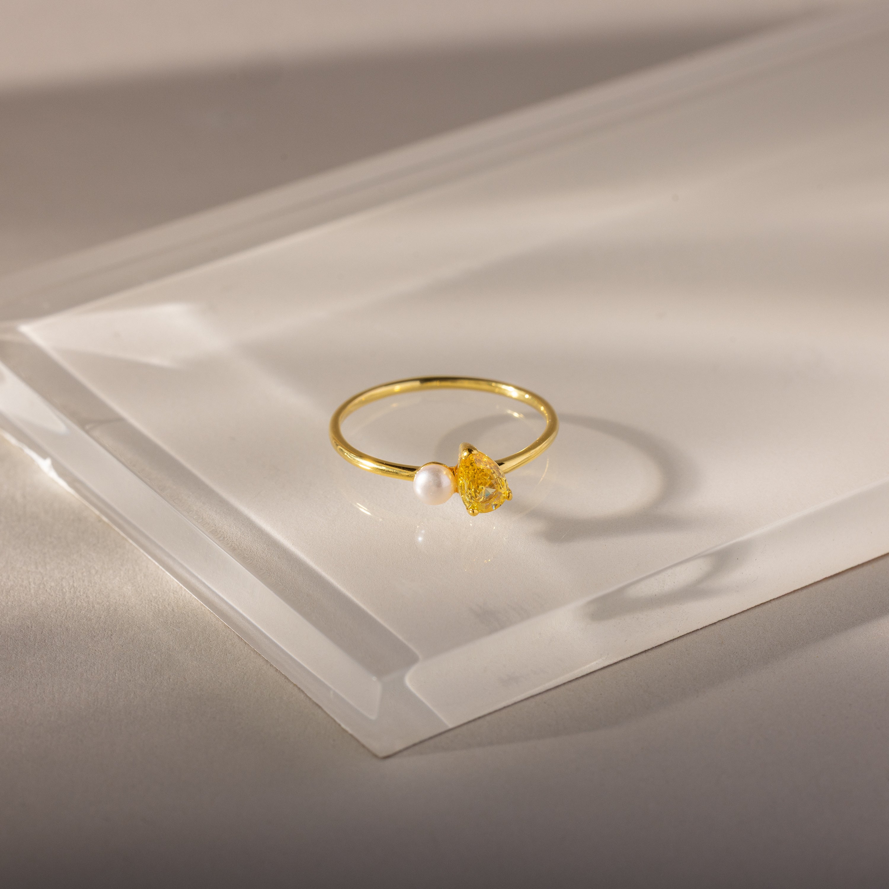 The Teardrop Topaz & Pearl Ring, featuring a yellow topaz and a white pearl, sits on a translucent surface, highlighting elegant jewelry perfect for any occasion.