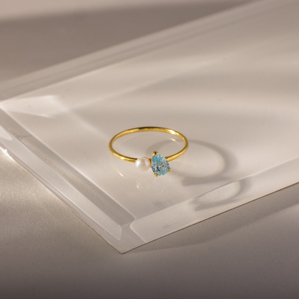The Teardrop Aquamarine & Pearl Ring features a blue aquamarine gemstone and a pearl in an elegant design, showcased on a clear, angled surface.