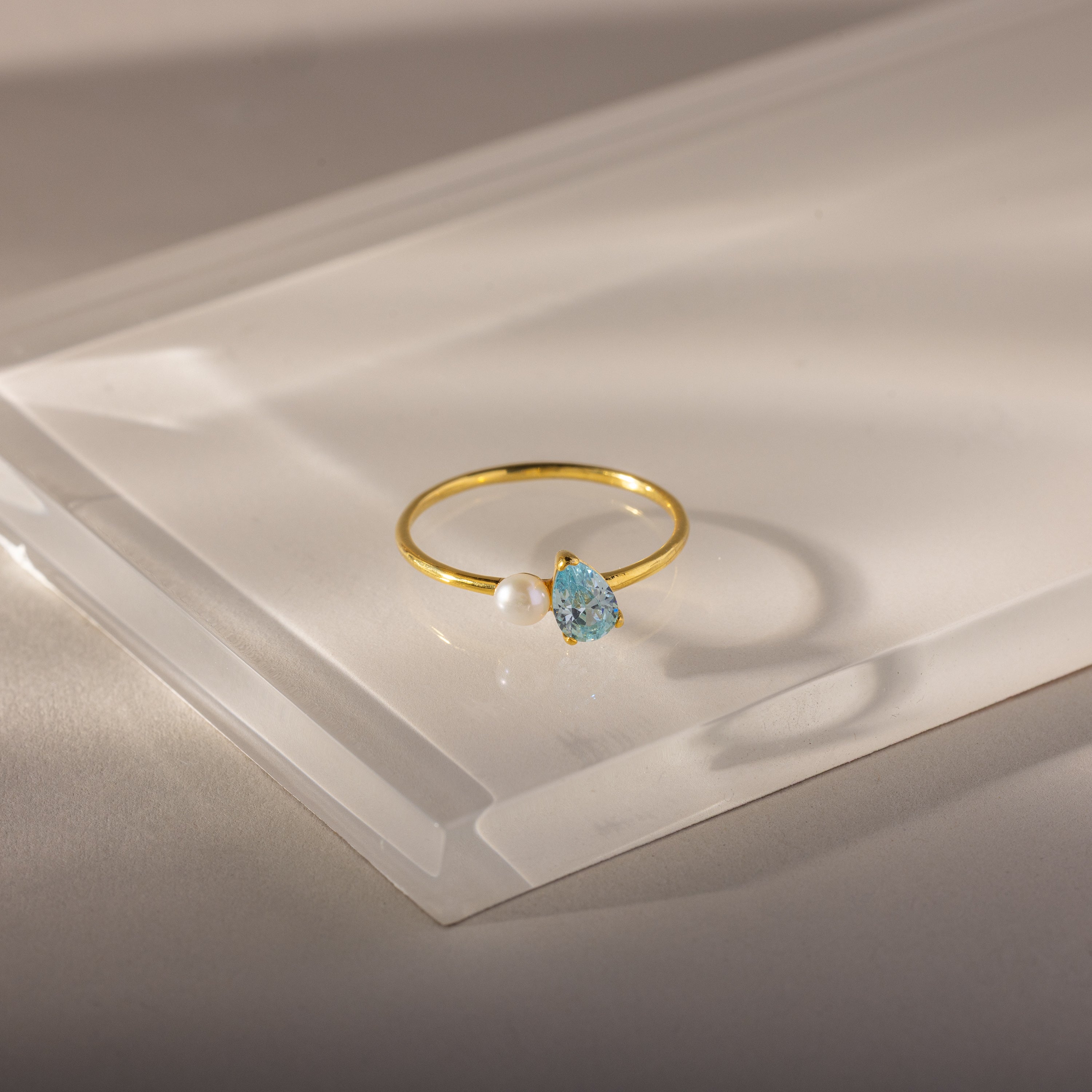 The Teardrop Aquamarine & Pearl Ring features a blue aquamarine gemstone and a pearl in an elegant design, showcased on a clear, angled surface.