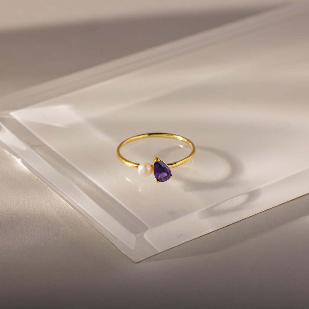 The Teardrop Amethyst & Pearl Ring features a gold band with a purple amethyst teardrop and a small white pearl, displayed on clear acrylic.