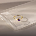 The Teardrop Amethyst & Pearl Ring features a gold band with a purple amethyst teardrop and a small white pearl, displayed on clear acrylic.