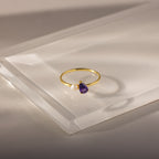 The Teardrop Amethyst & Pearl Ring features a gold band with a purple amethyst teardrop and a small white pearl, displayed on clear acrylic.