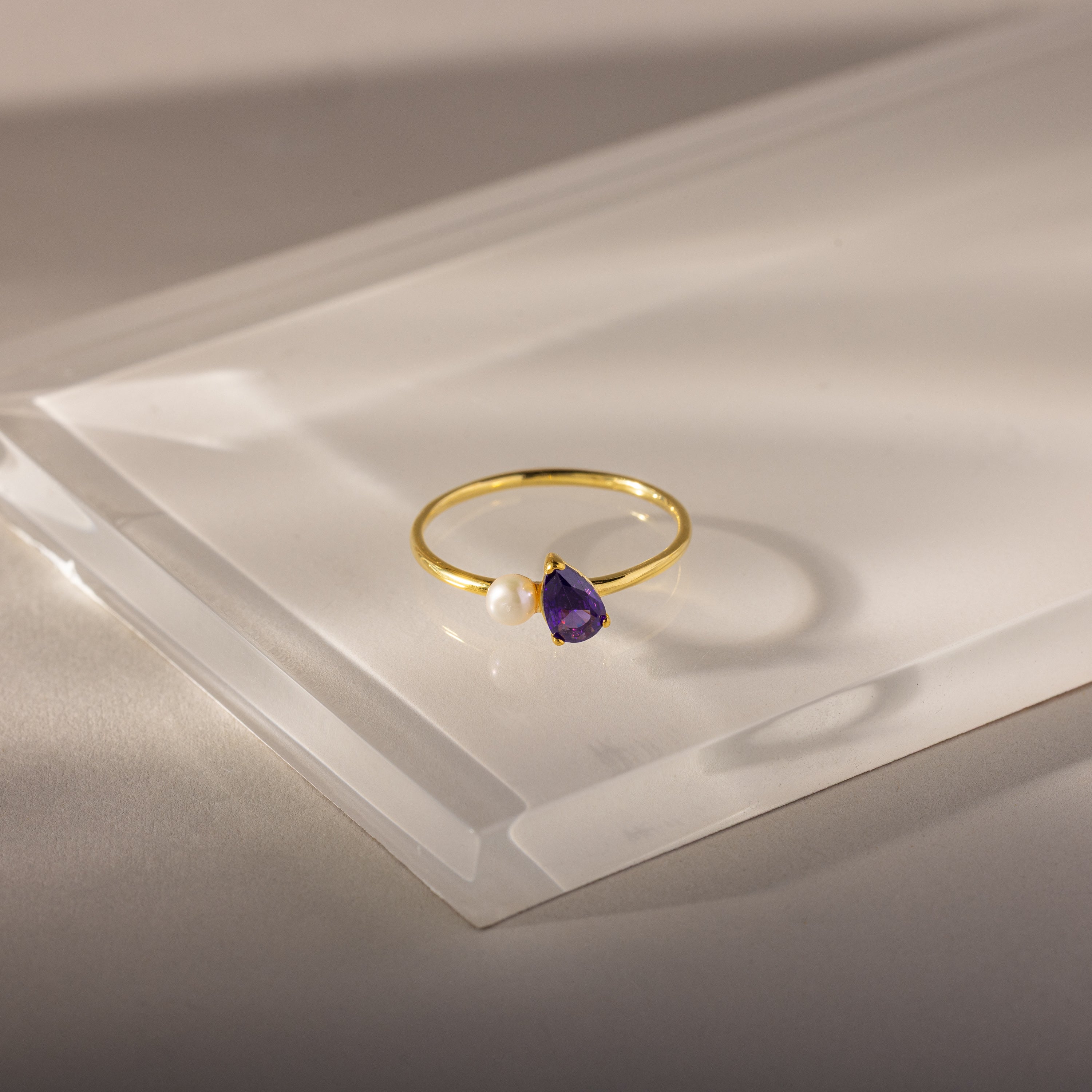The Teardrop Amethyst & Pearl Ring features a gold band with a purple amethyst teardrop and a small white pearl, displayed on clear acrylic.
