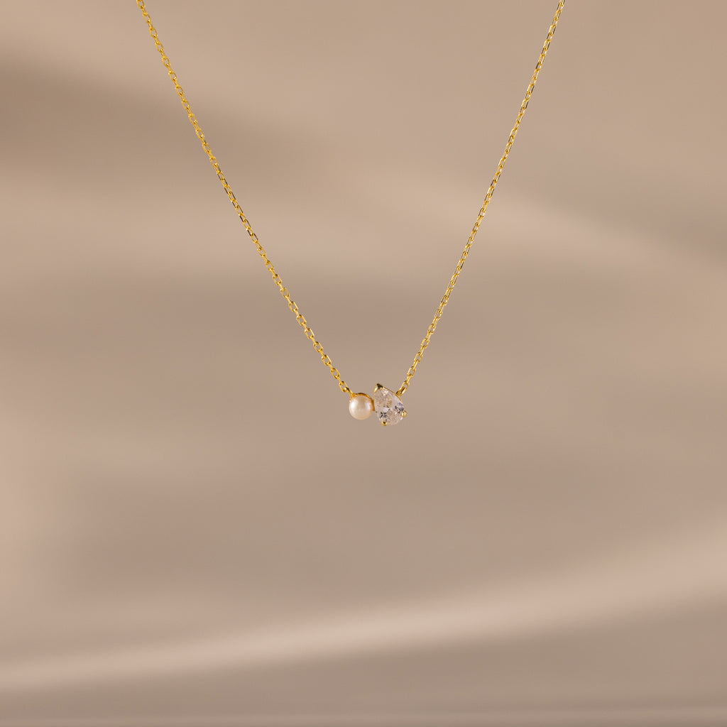 The Teardrop Diamond & Pearl Necklace features a delicate gold chain with a pearl and clear gemstone pendant, shown on a soft beige background—an elegant gift for any occasion.
