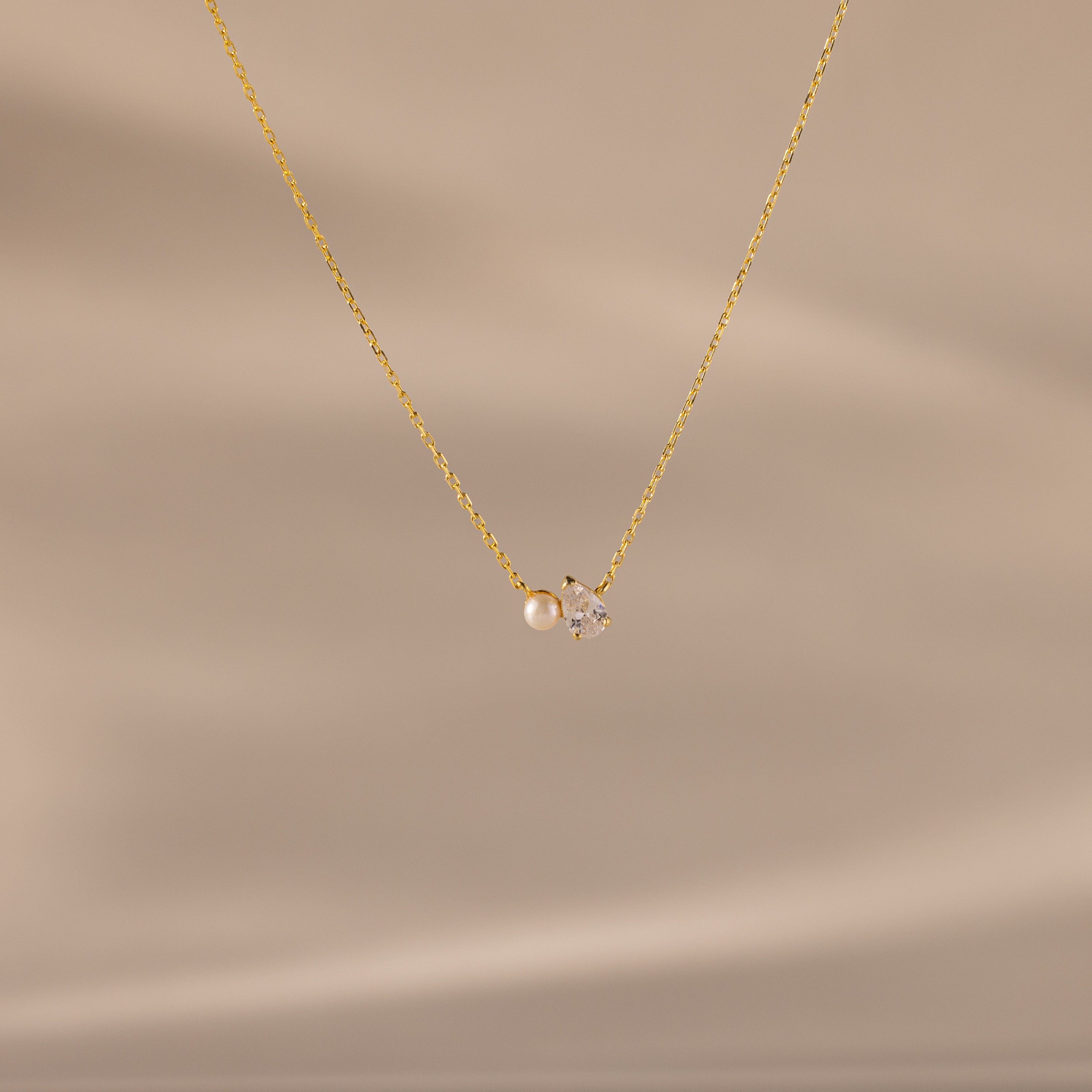 The Teardrop Diamond & Pearl Necklace features a delicate gold chain with a pearl and clear gemstone pendant, shown on a soft beige background—an elegant gift for any occasion.