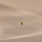 The Teardrop Peridot & Pearl Necklace, featuring a small green peridot and a white pearl pendant, rests on a soft beige background—a timeless gift for any occasion.