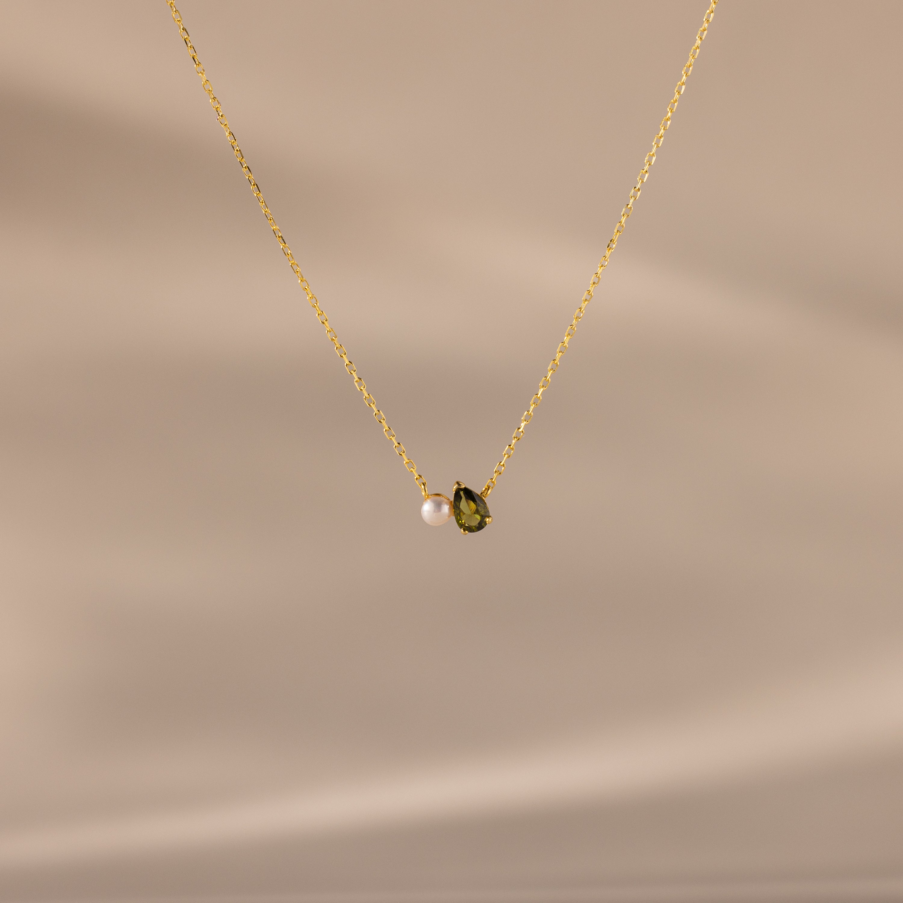 The Teardrop Peridot & Pearl Necklace, featuring a small green peridot and a white pearl pendant, rests on a soft beige background—a timeless gift for any occasion.