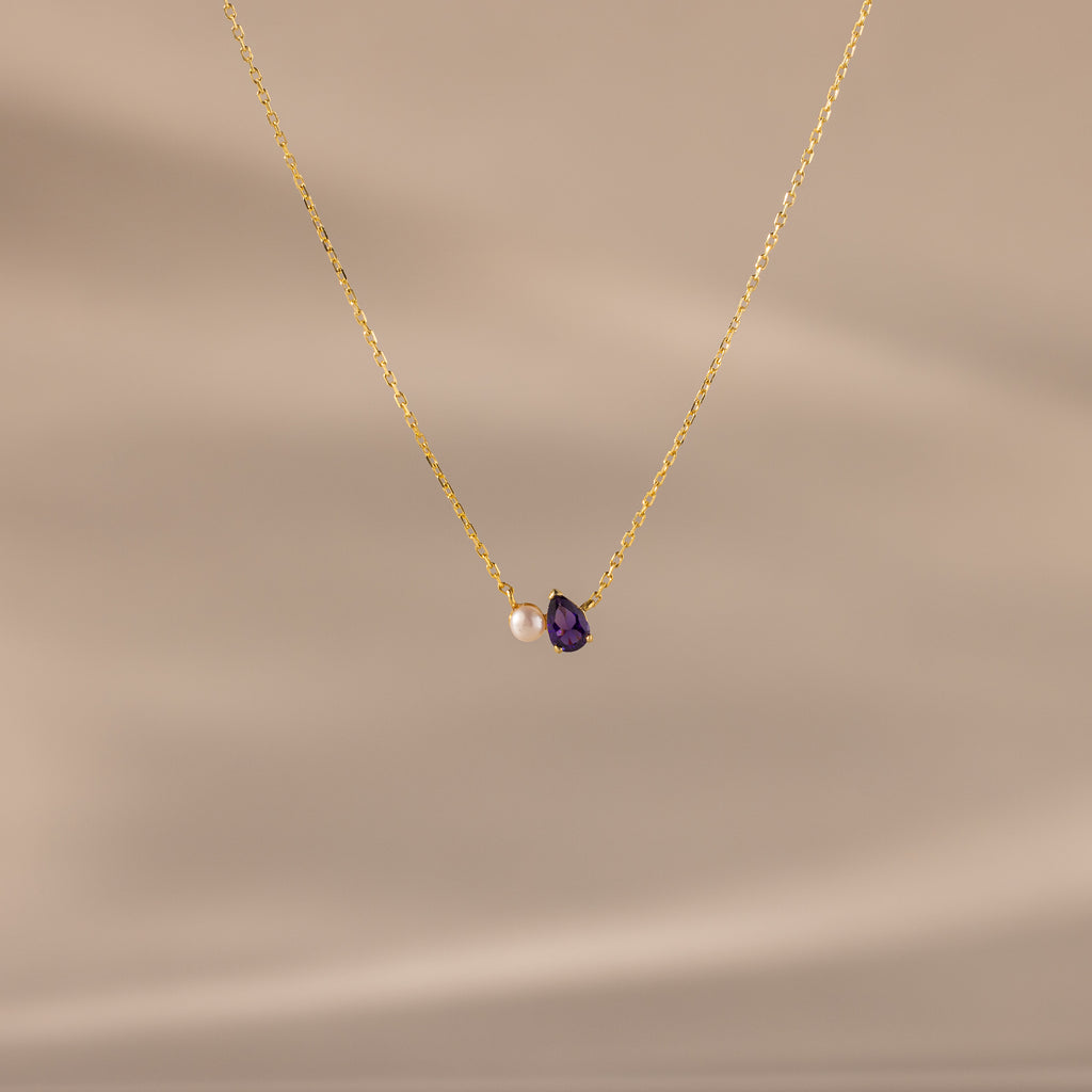 Teardrop Amethyst & Pearl Necklace featuring a gold chain, a delicate white pearl, and an amethyst teardrop pendant—an elegant gift for life's milestones, showcased against a soft beige background.