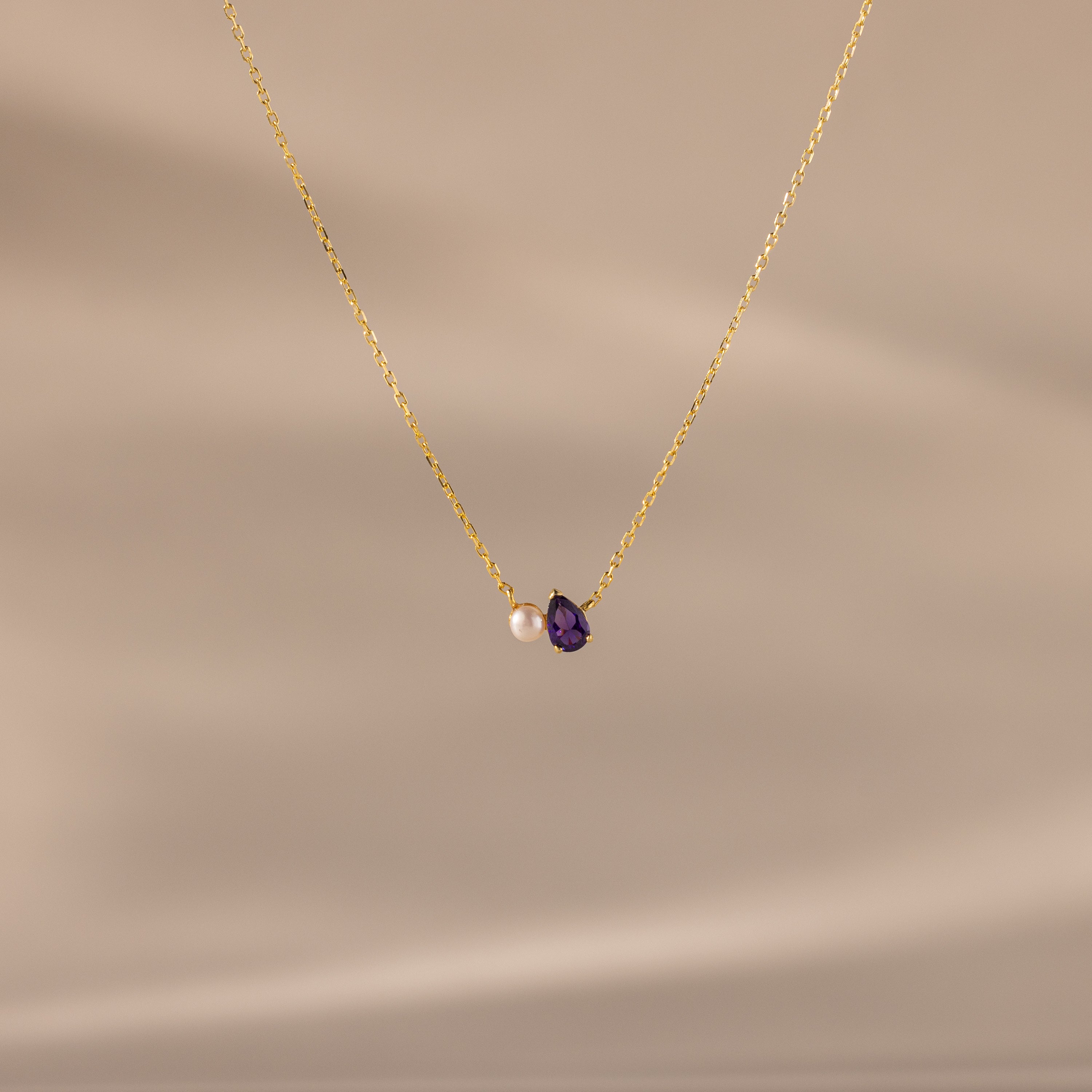 Teardrop Amethyst & Pearl Necklace featuring a gold chain, a delicate white pearl, and an amethyst teardrop pendant—an elegant gift for life's milestones, showcased against a soft beige background.