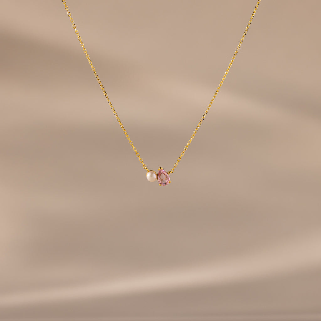 The Teardrop Pink Tourmaline & Pearl Necklace features a delicate gold teardrop pendant with a pink gemstone and pearl, set against a soft beige background—a timeless gift for any occasion.