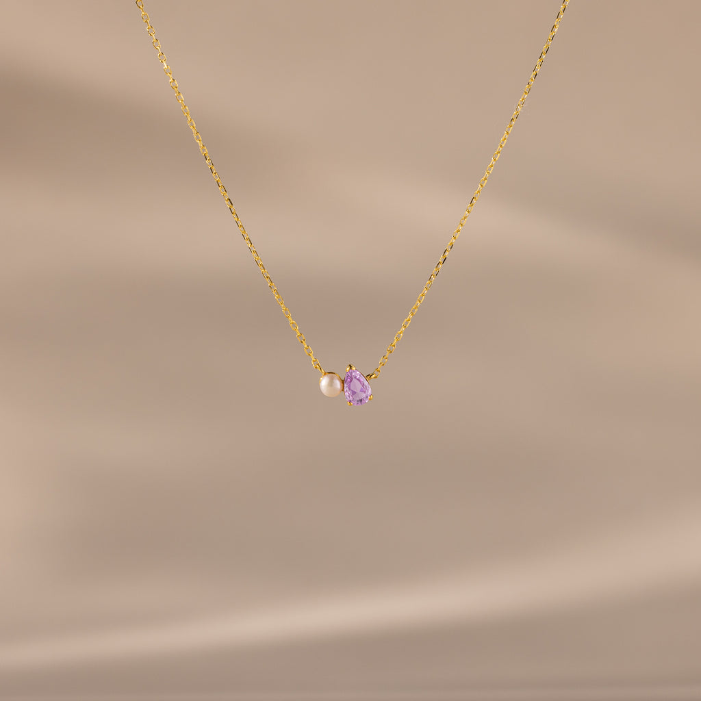 The Teardrop Alexandrite & Pearl Necklace features a gold chain with a small pearl and a purple teardrop gemstone pendant. A timeless gift for special occasions, shown here on a beige background.