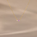 The Teardrop Alexandrite & Pearl Necklace features a gold chain with a small pearl and a purple teardrop gemstone pendant. A timeless gift for special occasions, shown here on a beige background.