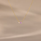 The Teardrop Alexandrite & Pearl Necklace features a gold chain with a small pearl and a purple teardrop gemstone pendant. A timeless gift for special occasions, shown here on a beige background.