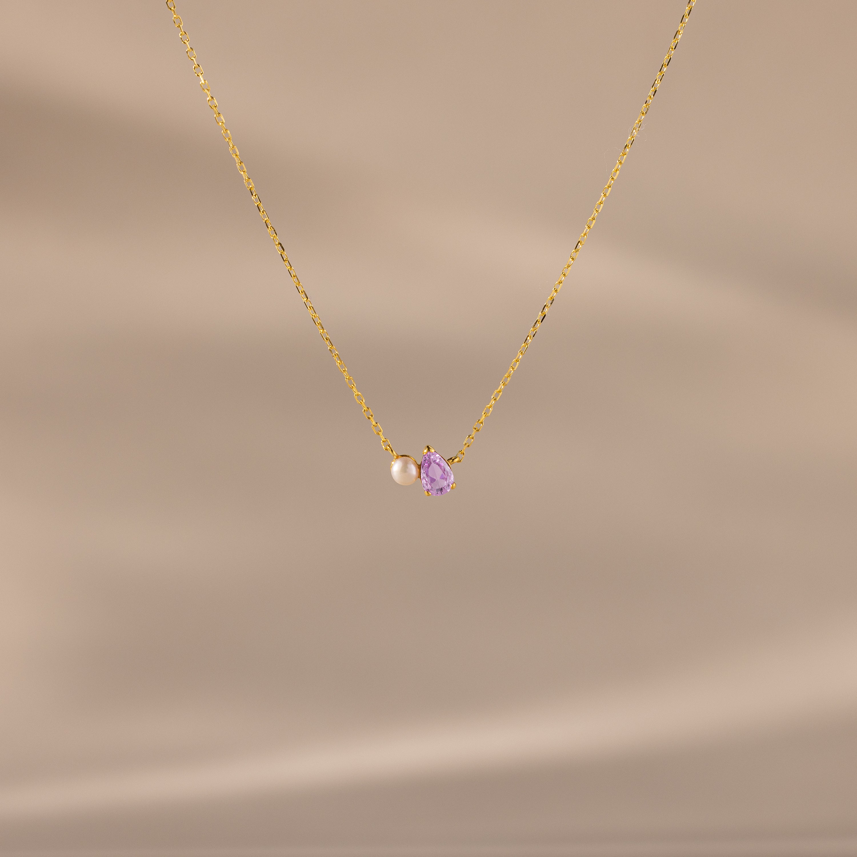 The Teardrop Alexandrite & Pearl Necklace features a gold chain with a small pearl and a purple teardrop gemstone pendant. A timeless gift for special occasions, shown here on a beige background.