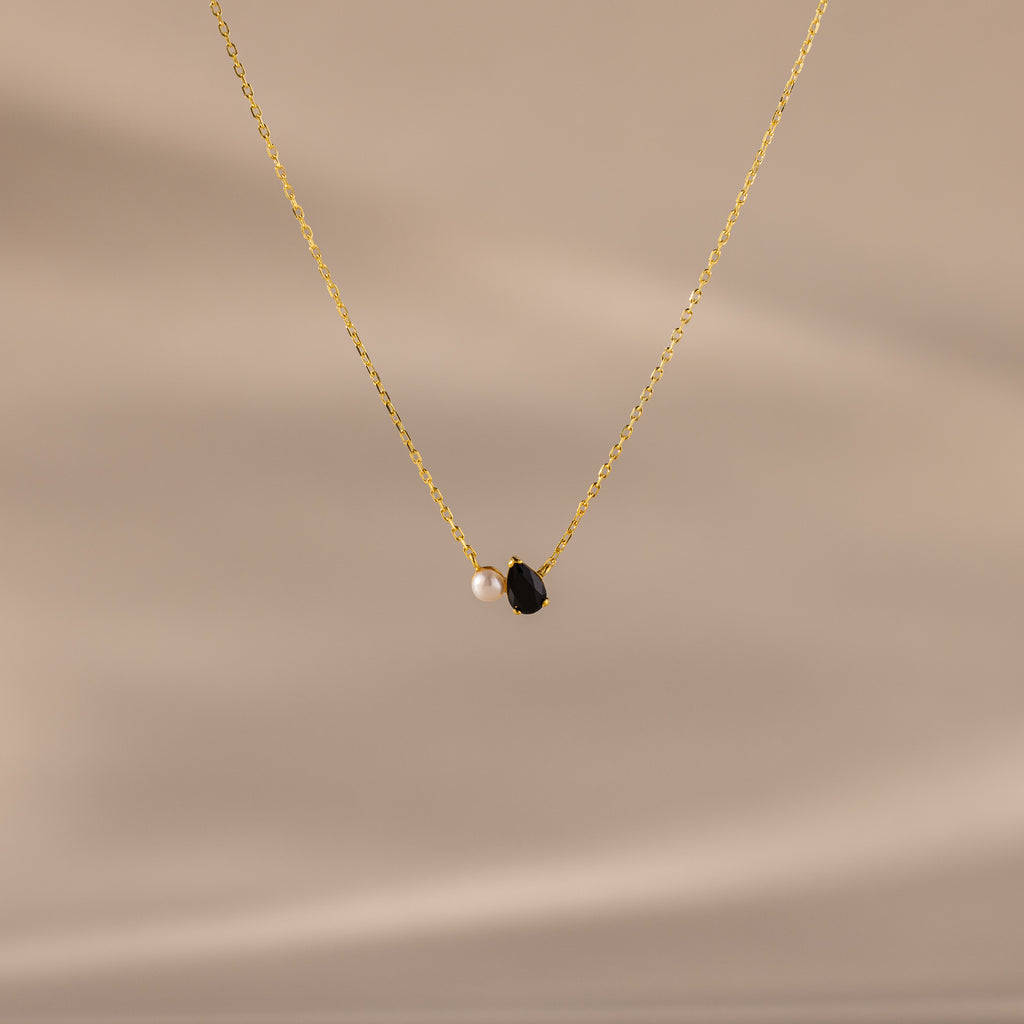 The Teardrop Onyx & Pearl Necklace features a gold chain with a delicate white pearl and black teardrop onyx pendant, displayed on a neutral beige background—an elegant, timeless gift for any occasion.