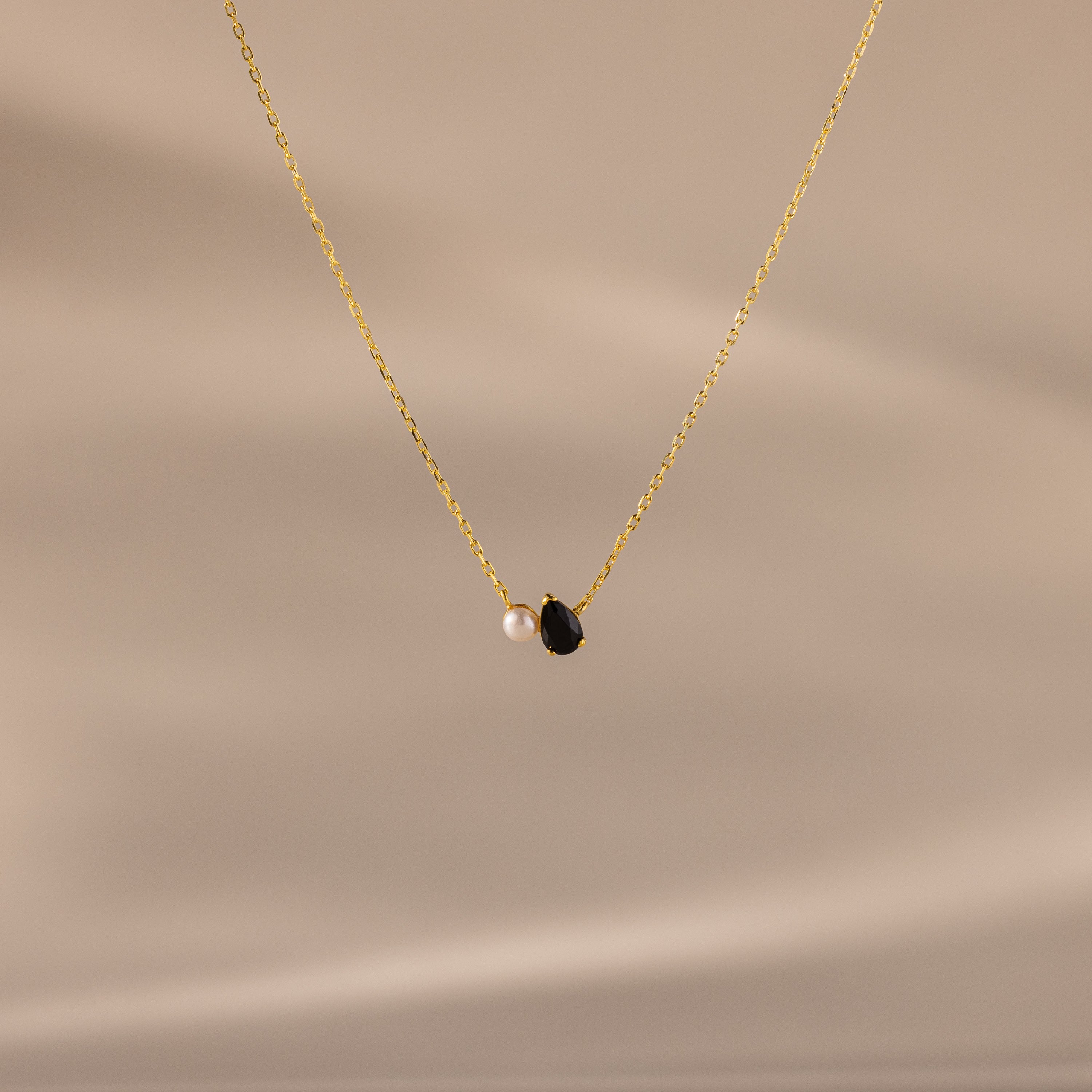 The Teardrop Onyx & Pearl Necklace features a gold chain with a delicate white pearl and black teardrop onyx pendant, displayed on a neutral beige background—an elegant, timeless gift for any occasion.