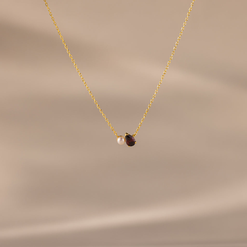 The Teardrop Garnet & Pearl Necklace features a small white pearl and a deep red garnet pendant on a delicate gold chain, making it an elegant milestone gift. Displayed on a beige background.
