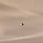 The Teardrop Garnet & Pearl Necklace features a small white pearl and a deep red garnet pendant on a delicate gold chain, making it an elegant milestone gift. Displayed on a beige background.