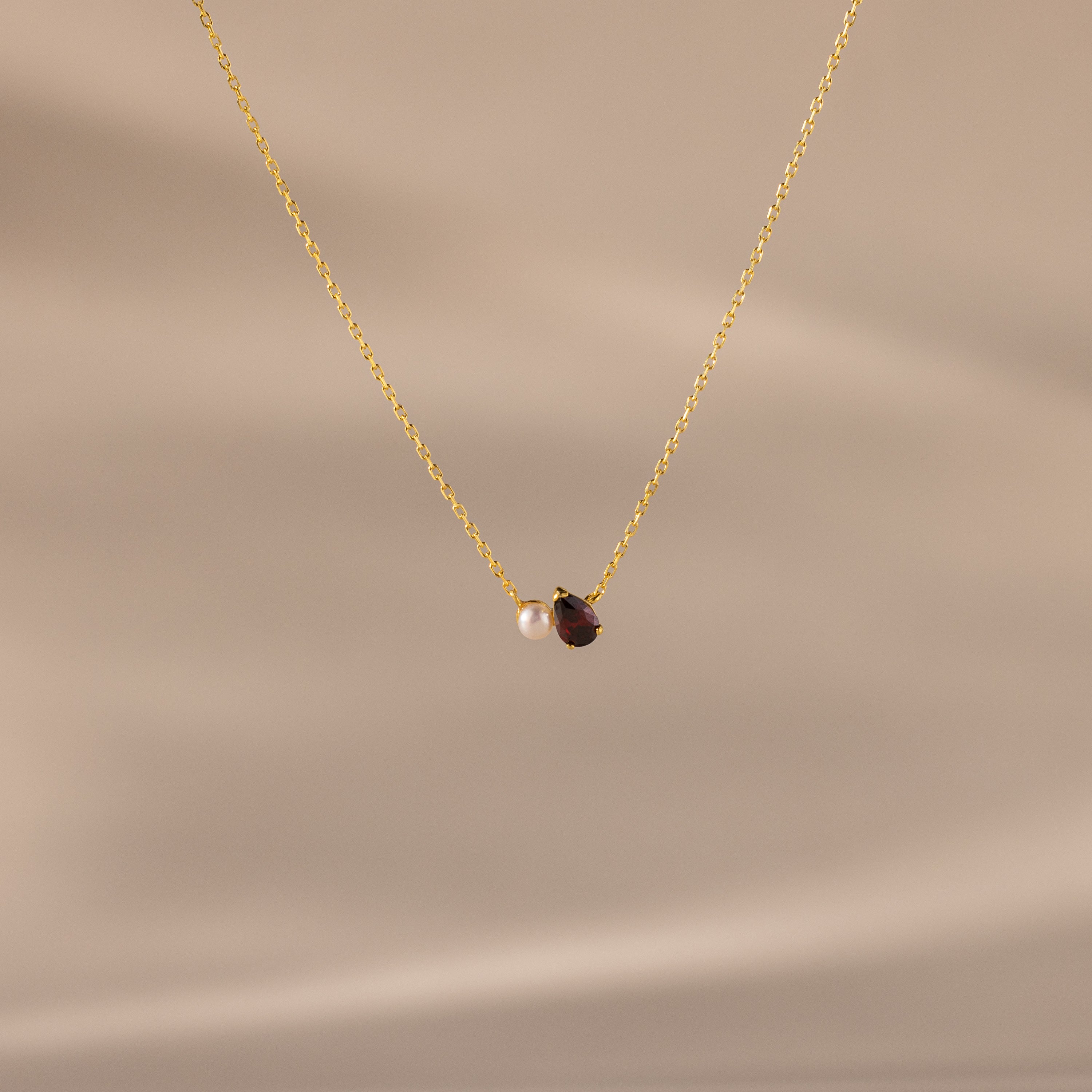 The Teardrop Garnet & Pearl Necklace features a small white pearl and a deep red garnet pendant on a delicate gold chain, making it an elegant milestone gift. Displayed on a beige background.